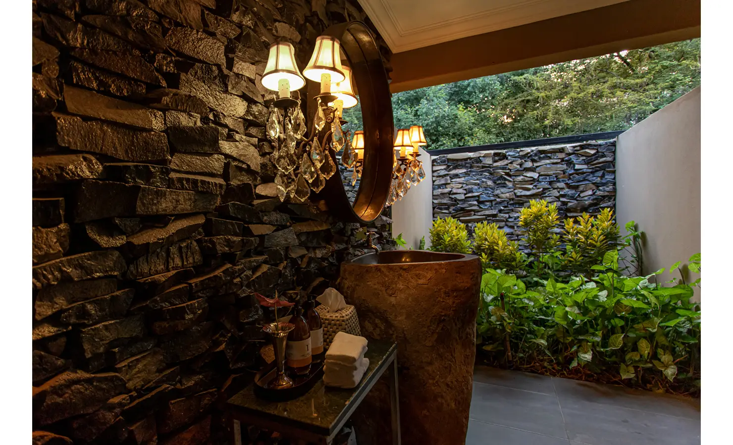 Perfect Hideaways: semi-open washroom with stacked-stone walls, rock-hewn basin and crystal wall sconces overlooking a planted courtyard — Mukwa River Lodge, Livingstone