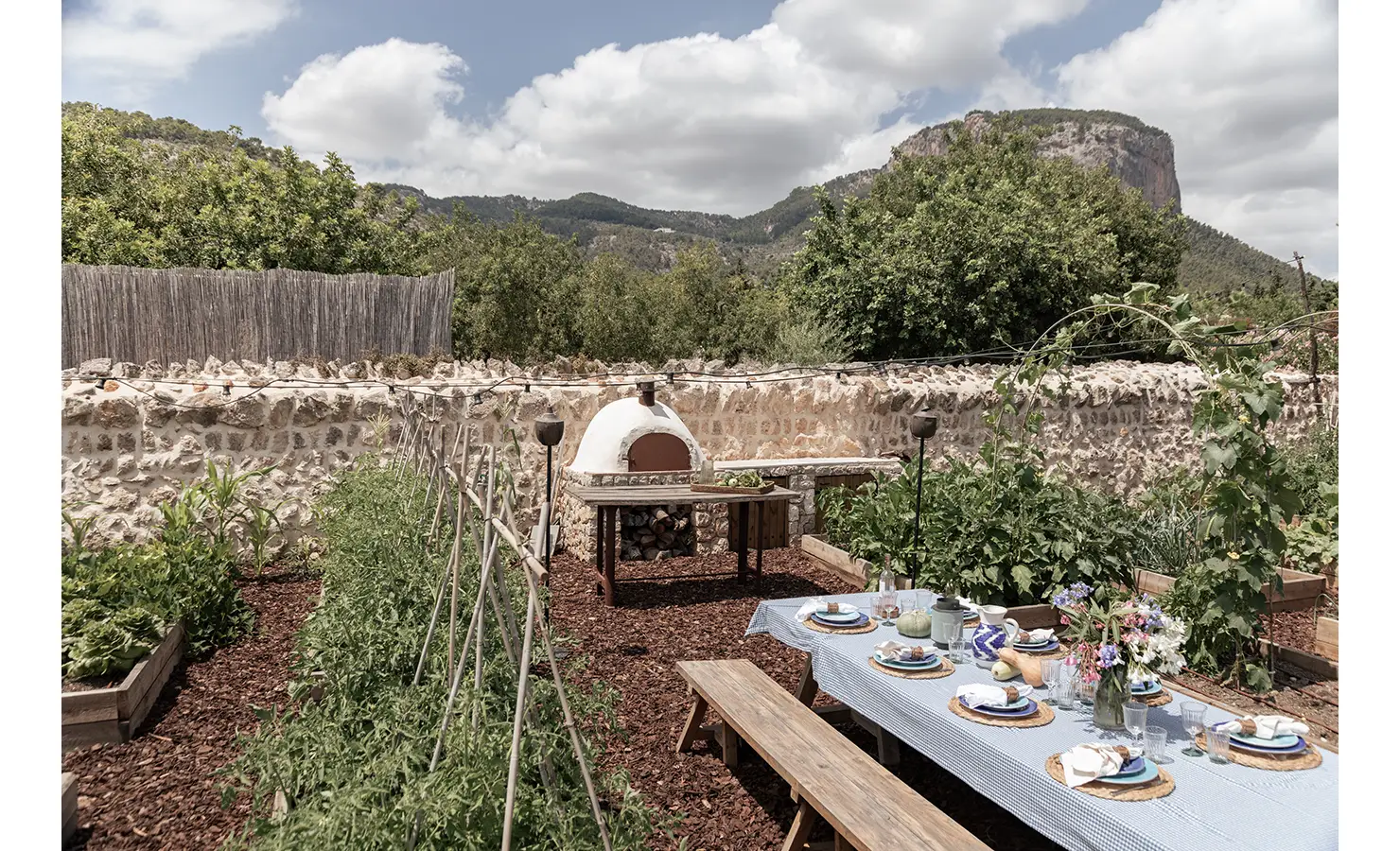 Perfect Hideaways: A picturesque vegetable garden bordered by stone walls and overlooked by dramatic cliffs, with a long outdoor dining table and a traditional wood-fired pizza oven nestled among verdant greenery. Finca Es Rafal, Alaró