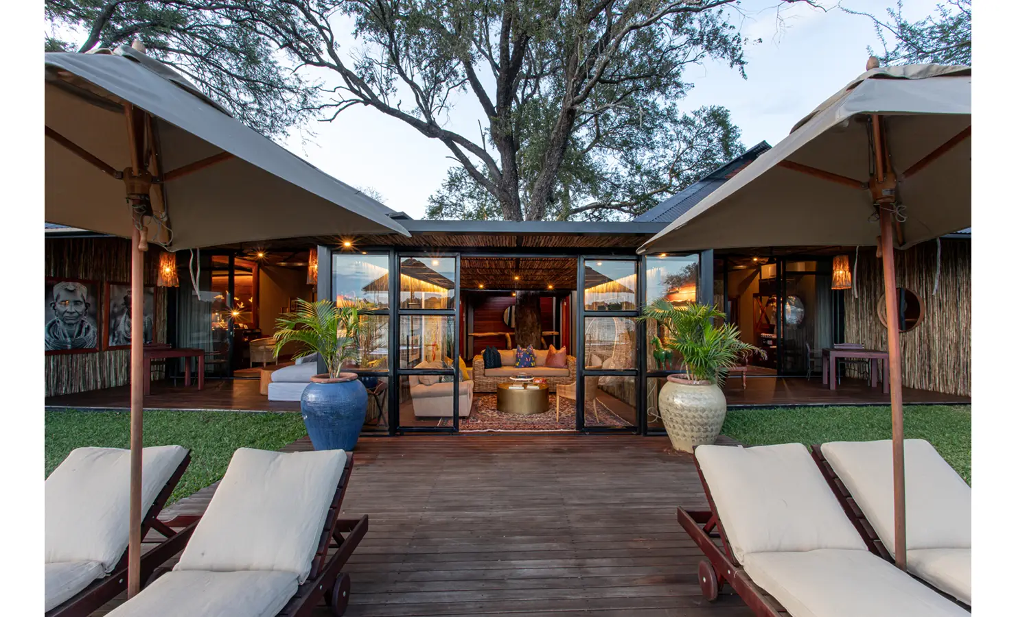 Perfect Hideaways: sun deck view of the main lodge with wide glass doors, canvas parasols, cushioned loungers and oversized ceramic urns — Mukwa River Lodge, Livingstone
