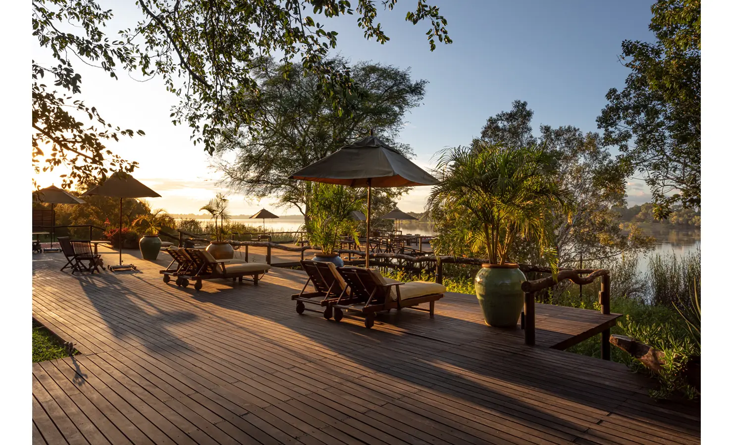 Perfect Hideaways: sunrise river deck with canvas-topped loungers, parasols and potted palms, shaded by trees and overlooking still water — Mukwa River Lodge, Livingstone