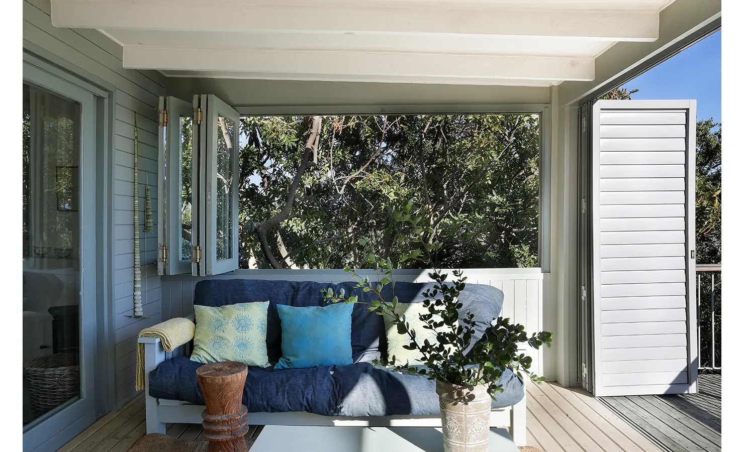 Perfect Hideaways: Cosy deck corner with navy-cushioned bench, sea-green scatter cushions, open shutters and a potted shrub under the eaves — tranquil treetop nook, Amongst The Trees, Scarborough.
