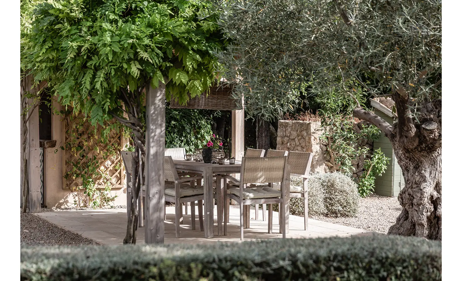 Perfect Hideaways: A shaded al fresco dining area beneath a leafy pergola, with pale timber chairs and a rustic table set against an olive grove and old stone walls. Finca Es Rafal, Alaró