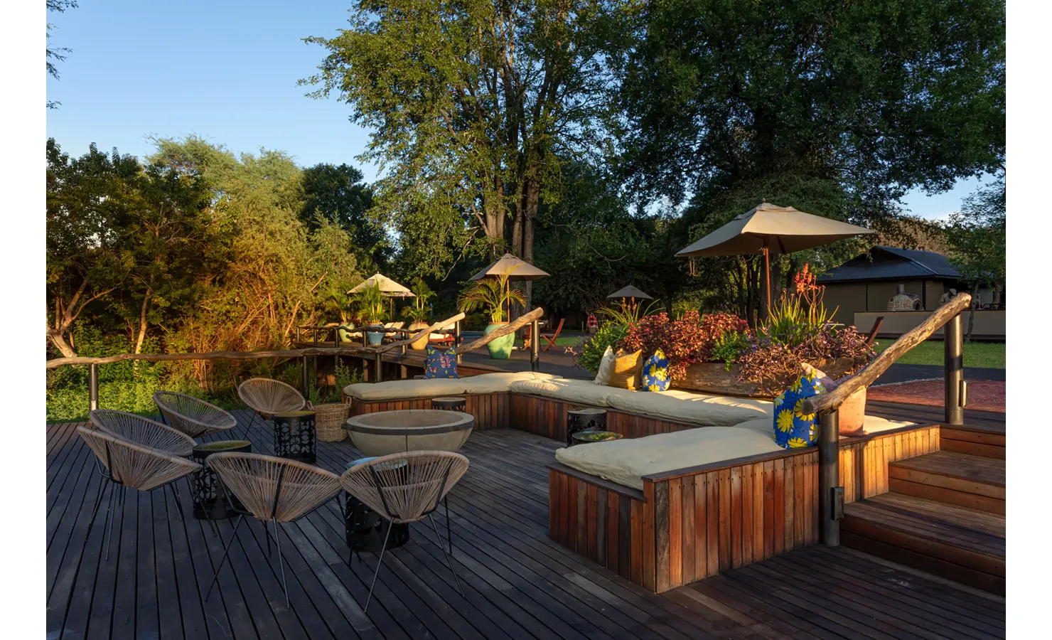 Perfect Hideaways: tiered timber deck with built-in cushioned seating, rope-weave chairs, round fire bowl, parasols and lush planter boxes — Mukwa River Lodge, Livingstone