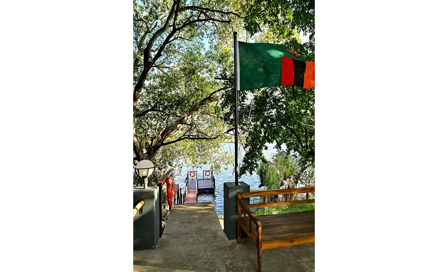 Perfect Hideaways: Pathway leading to a small riverside jetty with the Zambian flag fluttering above, shaded by large overhanging trees, Tanyati Villa, Livingstone
