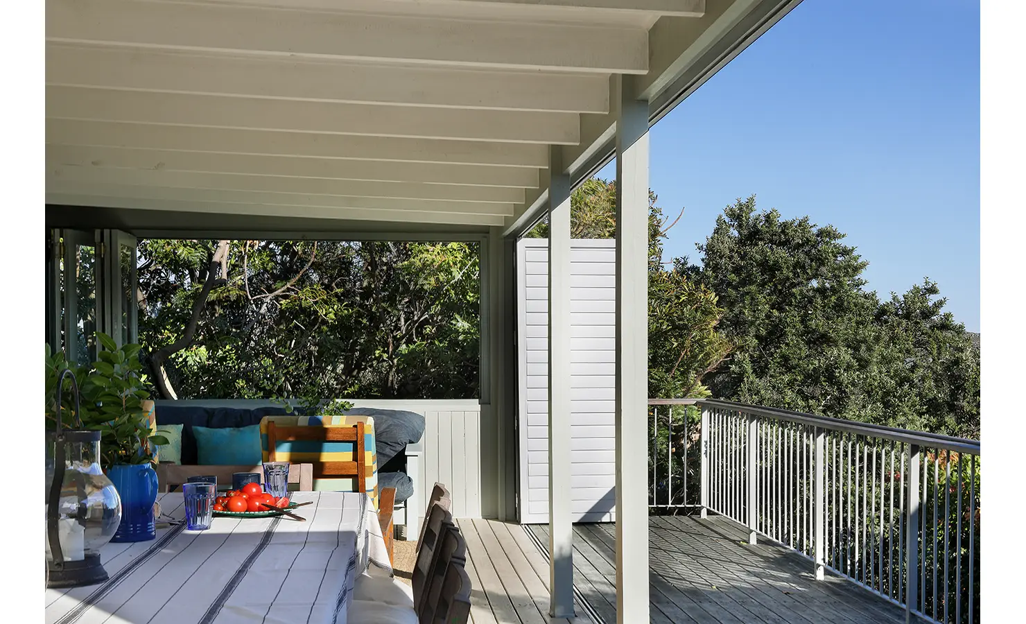 Perfect Hideaways: Long covered deck set with a striped-cloth table, blue glassware and tomatoes, leading to a cushioned daybed and leafy outlook — generous outdoor dining, Amongst The Trees, Scarborough.