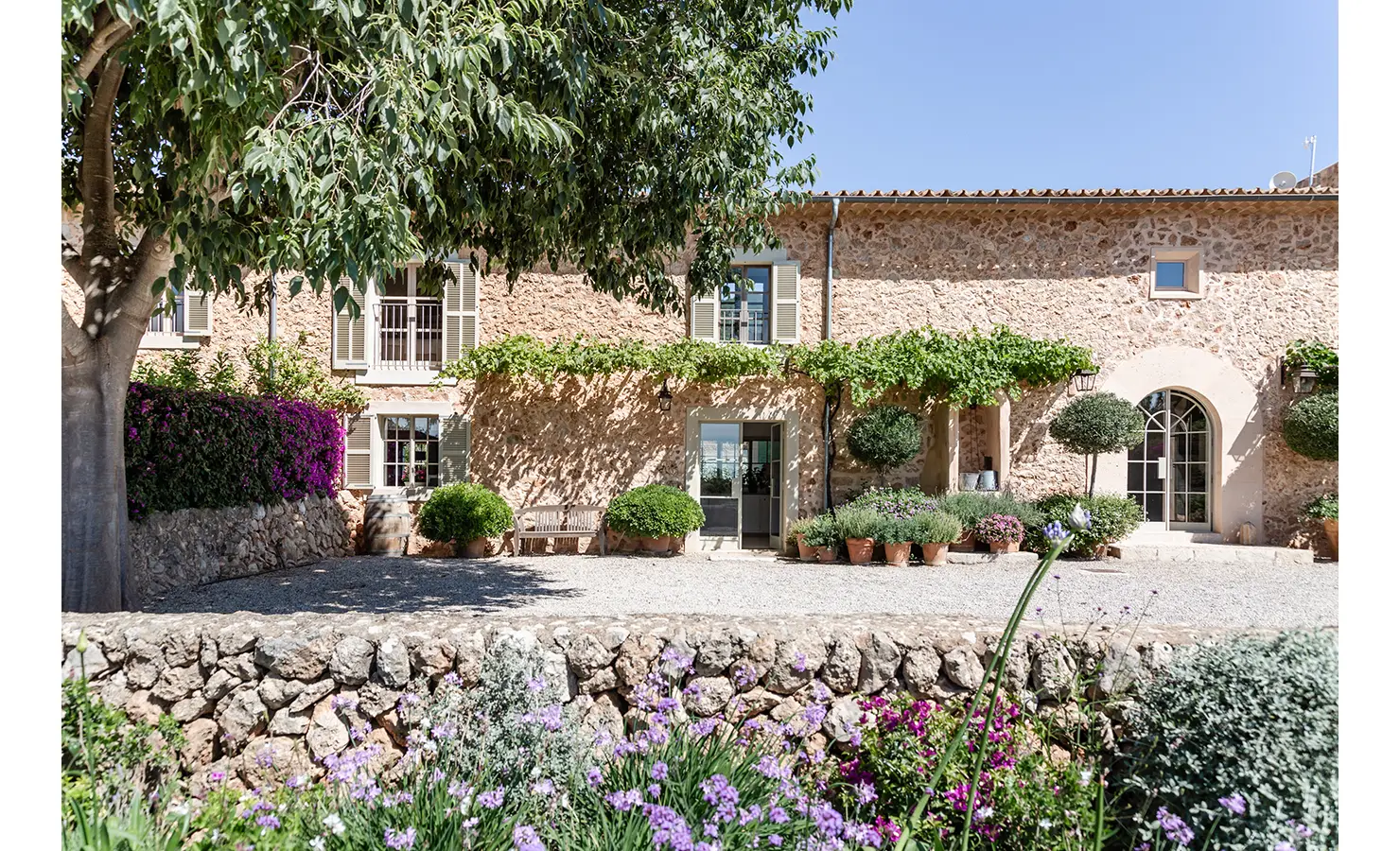 Perfect Hideaways: The entrance courtyard of a traditional Mallorcan finca with a stone façade, climbing vines, potted lavender and trees, framed by bougainvillea and Mediterranean sunshine. Finca Es Rafal, Alaró