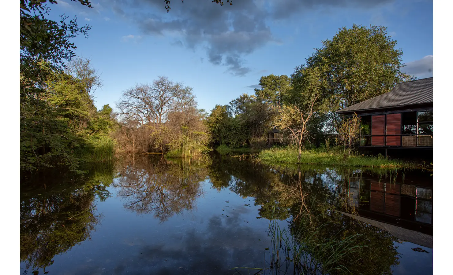 Perfect Hideaways: tranquil lagoon reflecting riverine trees with a timber-clad lodge pavilion on the bank under a wide blue sky — Mukwa River Lodge, Livingstone