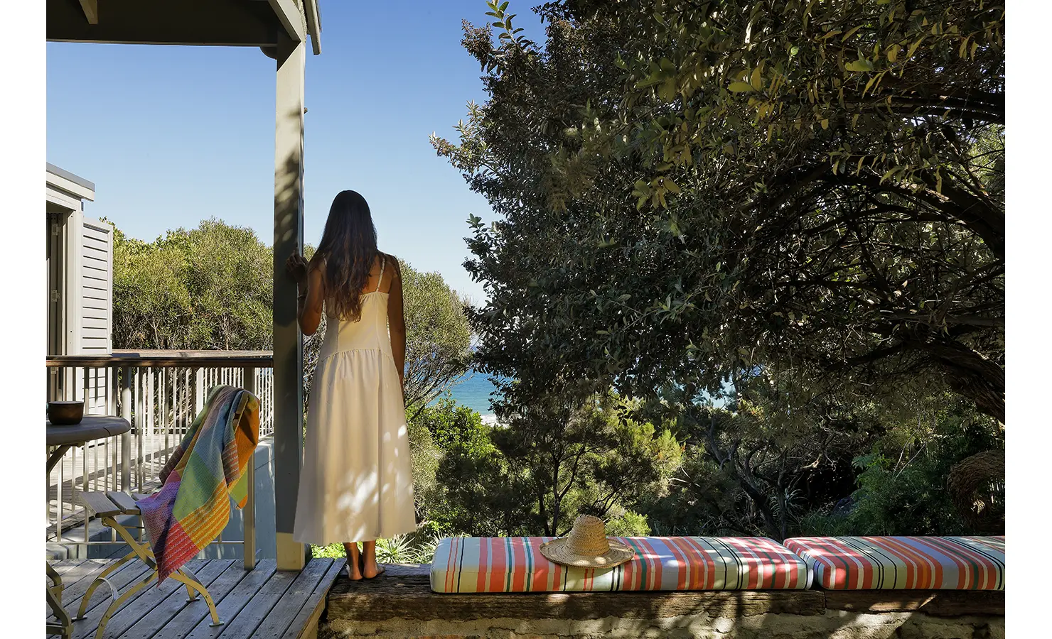 Perfect Hideaways: Guest in a white sundress gazing through trees to the ocean, striped bench pad with a straw hat catching dappled light — a quiet coastal moment, Amongst The Trees, Scarborough.