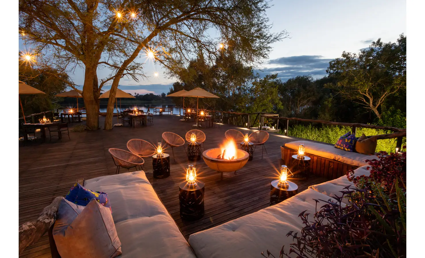 Perfect Hideaways: lantern-lit deck at dusk with a glowing firepit, string lights in the trees and relaxed riverside dining — Mukwa River Lodge, Livingstone