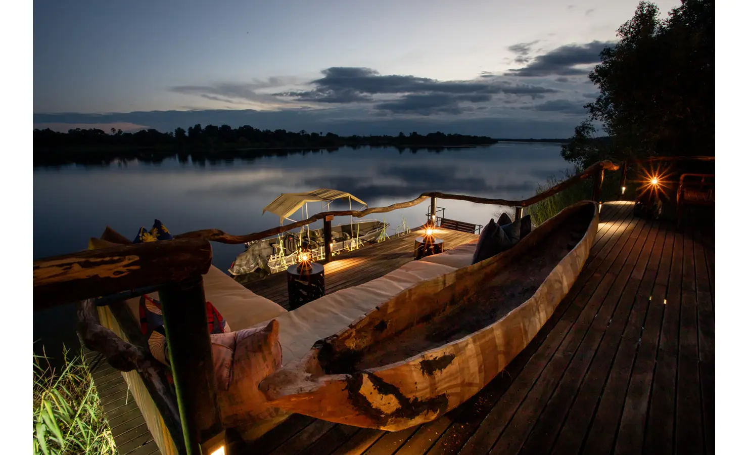 Perfect Hideaways: evening river deck with cushioned daybeds, sculptural dugout canoe and lanterns above a moored boat at the jetty — Mukwa River Lodge, Livingstone