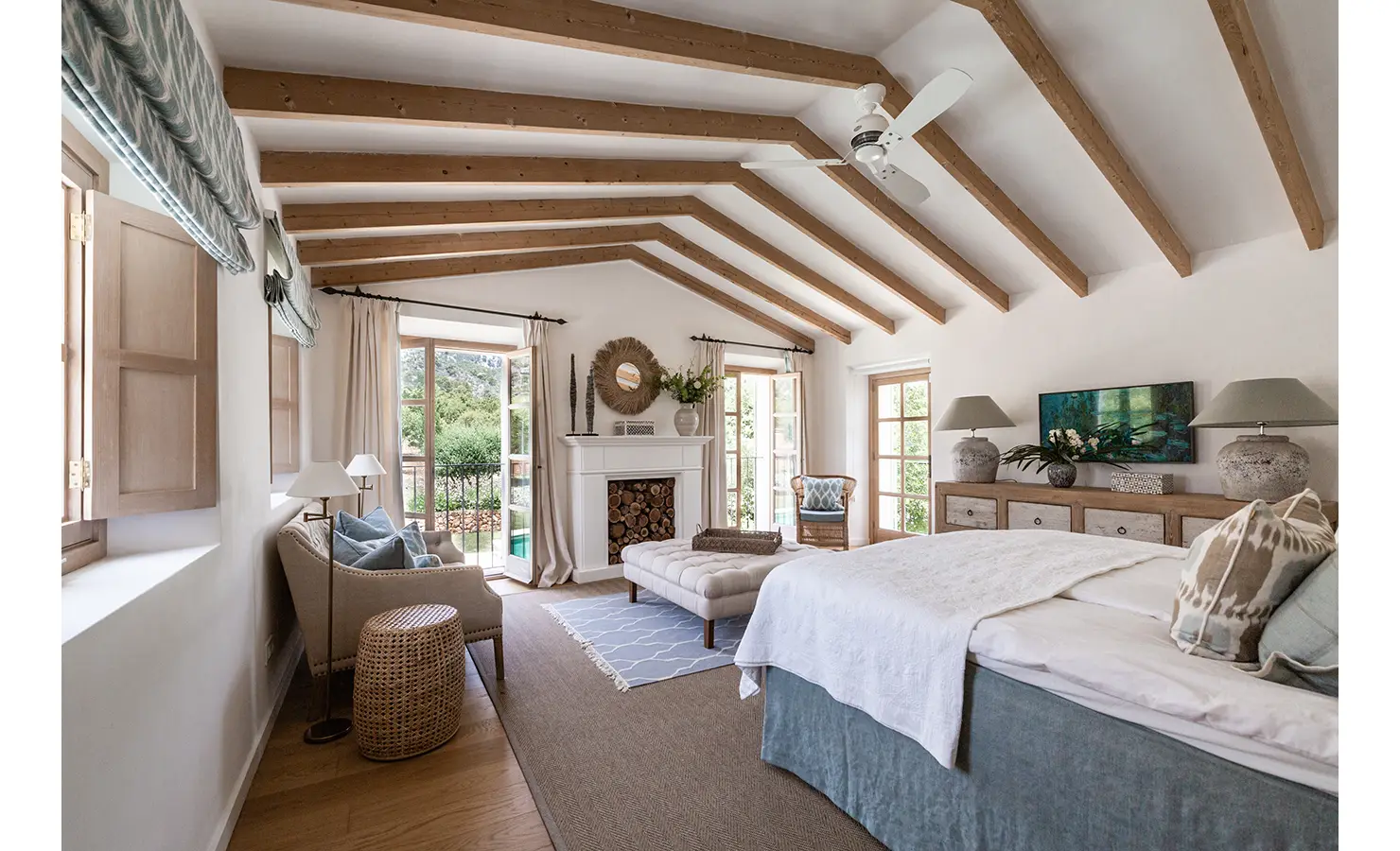 Perfect Hideaways: A dreamy bedroom with exposed beams, French doors on both sides, and soft neutral tones, complete with a fireplace and garden views from every angle. Finca Es Rafal, Alaró