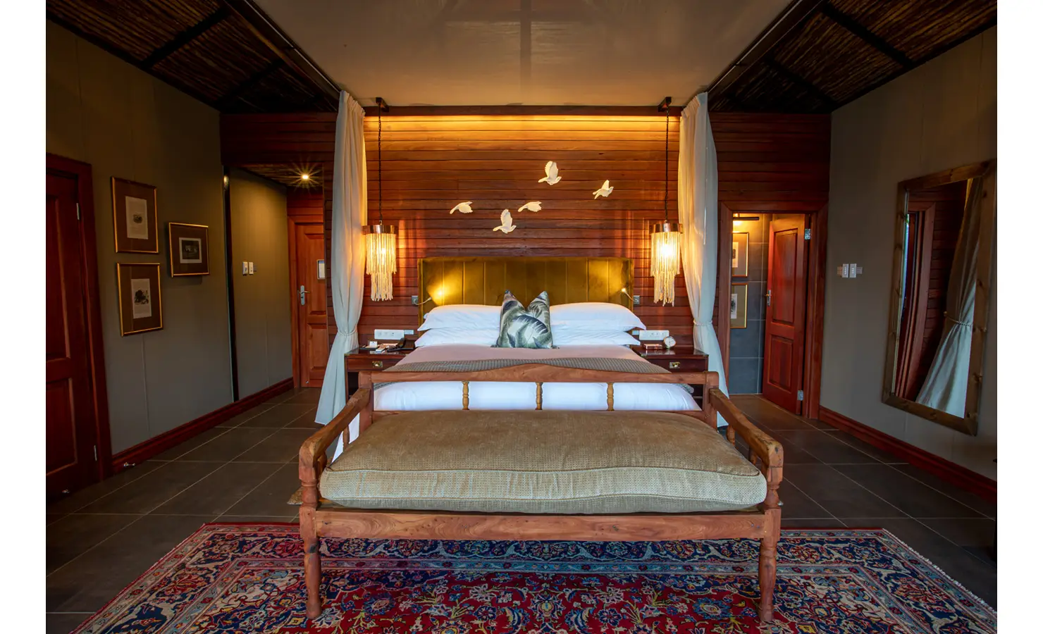 Perfect Hideaways: warmly lit bedroom with canopied bed, velvet headboard, pendant fringe lamps, bench and Persian rug — Mukwa River Lodge, Livingstone