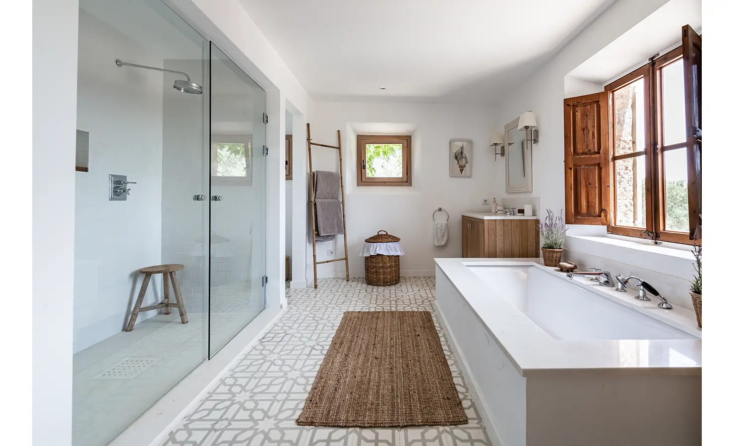 Perfect Hideaways: A bright and spacious bathroom with patterned floor tiles, a walk-in glass shower, freestanding bathtub, and natural wood accents, illuminated by shuttered windows. Finca Es Rafal, Alaró