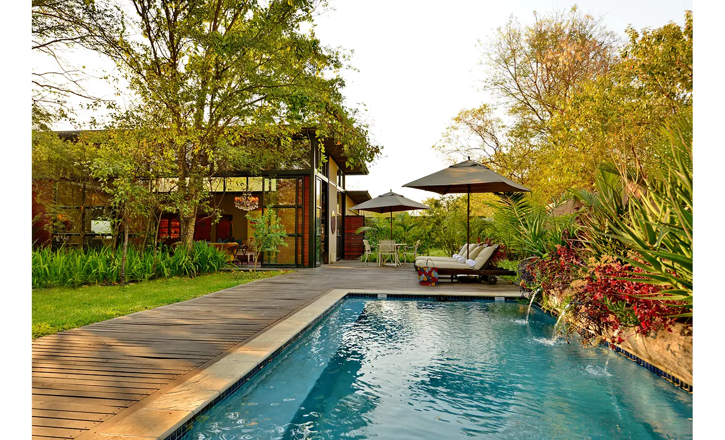 Perfect Hideaways: garden pool terrace with water spouts, sun loungers beneath parasols and a glass-fronted pavilion amid tropical planting — Mukwa River Lodge, Livingstone