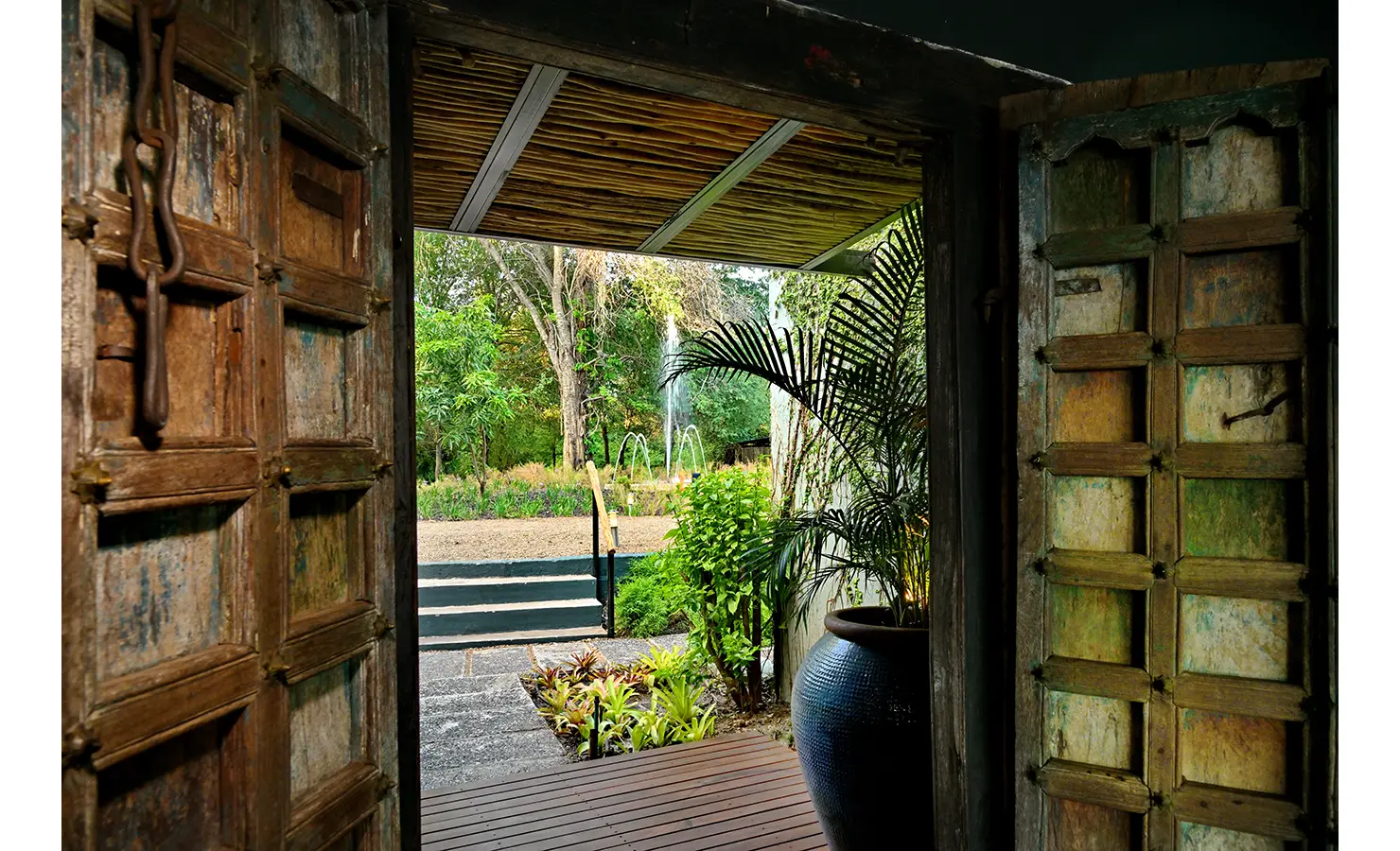Perfect Hideaways: View from a rustic carved wooden doorway onto lush gardens with a central fountain feature, Tanyati Villa, Livingstone