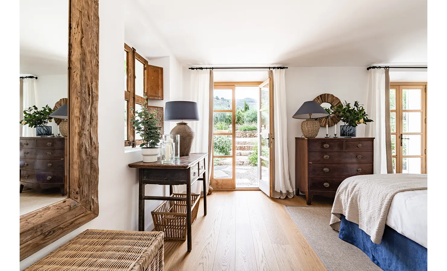 Perfect Hideaways: A charming bedroom corner with rustic wood furnishings, terracotta lamps, and soft natural light flowing in through French doors opening onto a garden path. Finca Es Rafal, Alaró
