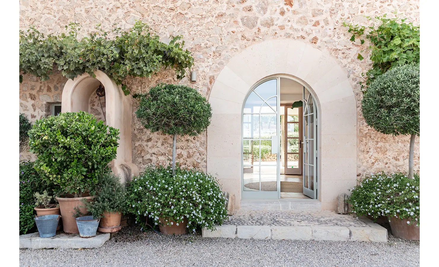 Perfect Hideaways: A graceful arched entrance in a stone wall façade framed by vines and clipped shrubs, welcoming guests through elegant glass doors into a serene interior. Finca Es Rafal, Alaró