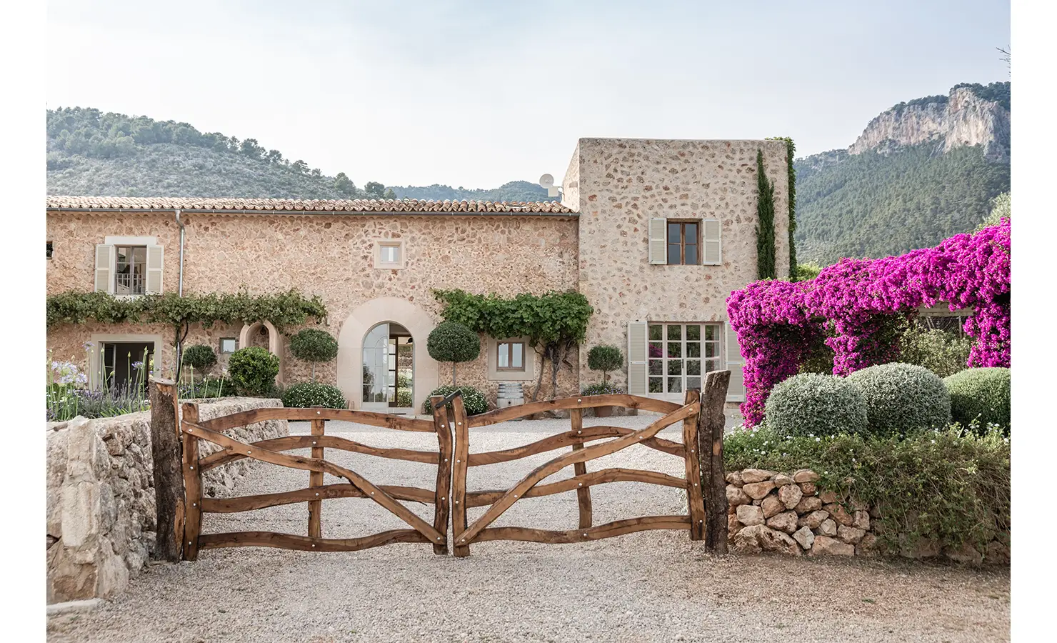 Perfect Hideaways: A rustic double-gated entrance leading to a honey-coloured stone finca, framed by manicured topiary and vibrant pink bougainvillea spilling over the courtyard pergola. Finca Es Rafal, Alaró