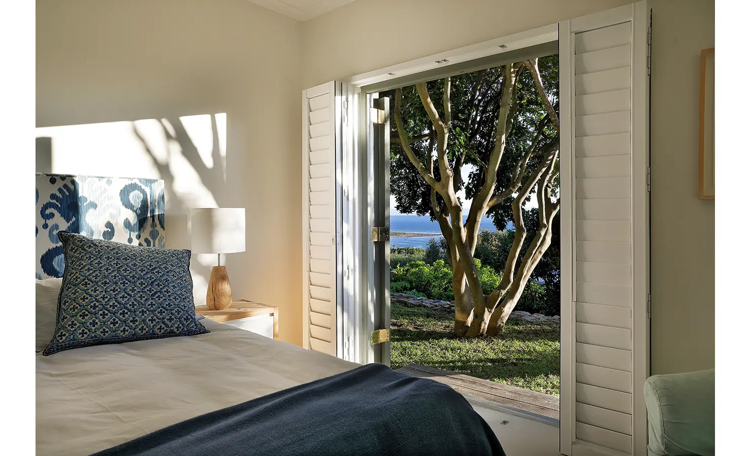 Perfect Hideaways: Bedroom in blue tones with ikat headboard and timber lamp; shutters open to a tree-framed ocean view — wake up to the coast, Amongst The Trees, Scarborough.