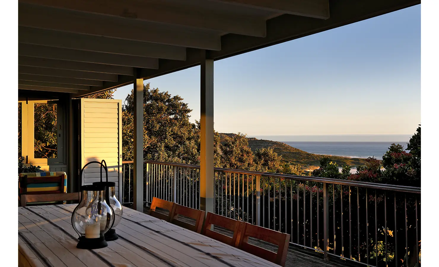 Perfect Hideaways: Long verandah table set with lanterns at golden hour, warm light on coastal hills and the bay beyond — sundowner spot, Amongst The Trees, Scarborough.
