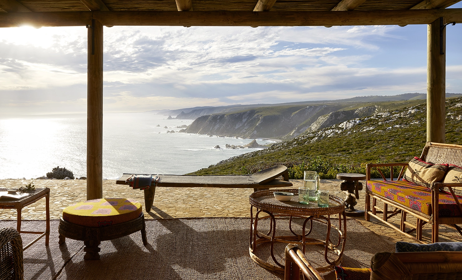 Perfect Hideaways: A rustic covered terrace with woven rattan furniture overlooks a dramatic coastline, where cliffs plunge into the ocean under a luminous sky, creating a serene and secluded retreat. Fynbos Fort, Plettenberg Bay.