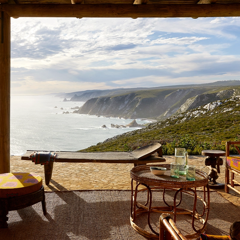 Perfect Hideaways: A rustic covered terrace with woven rattan furniture overlooks a dramatic coastline, where cliffs plunge into the ocean under a luminous sky, creating a serene and secluded retreat. Fynbos Fort, Plettenberg Bay.