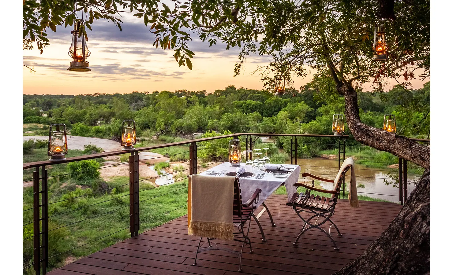 Perfect Hideaways: A romantic table for two is set on a wooden deck overlooking the Sand River, with vintage lanterns casting a golden glow beneath the canopy of a leafy tree at sunset. Dulini, Sabi Sand Nature Reserve