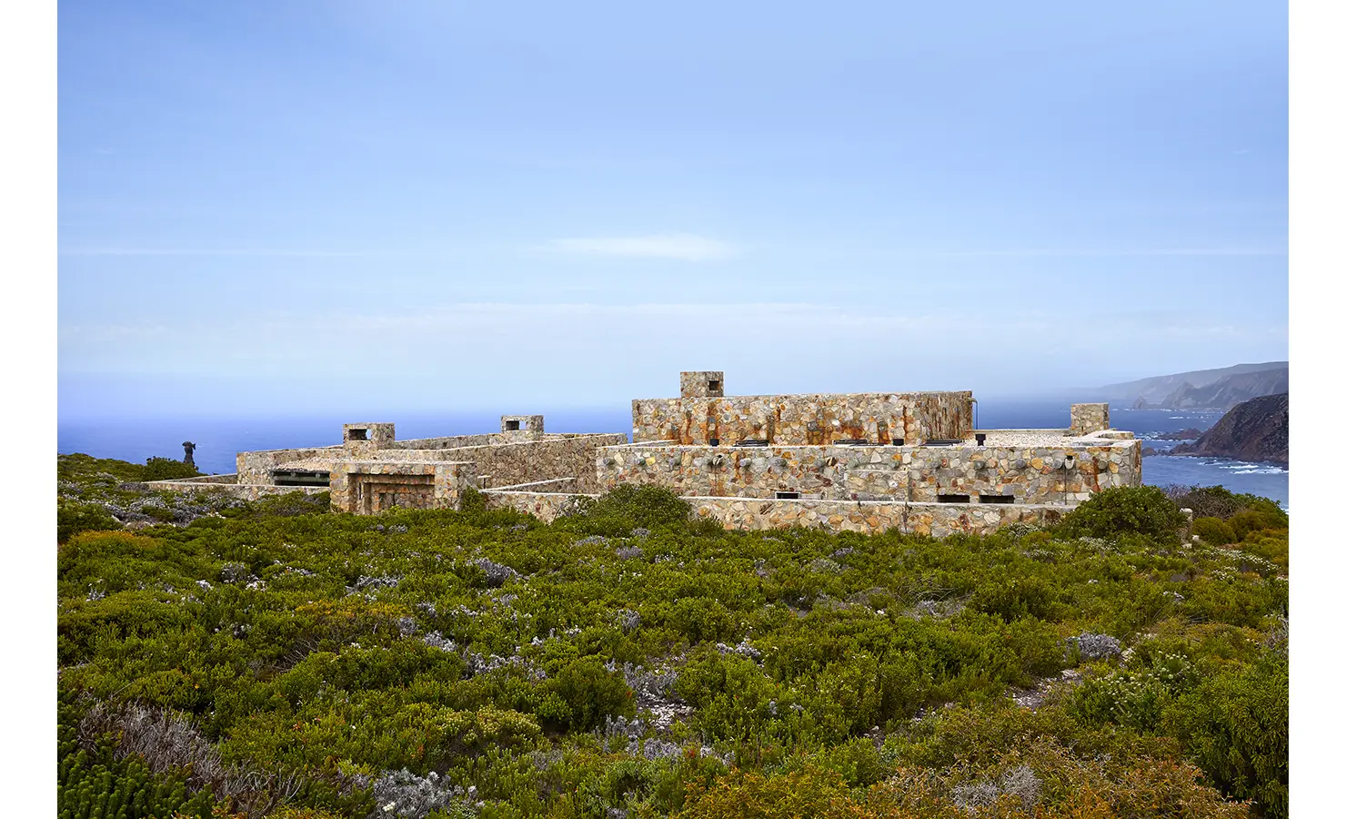 Perfect Hideaways: A striking stone-clad fort-like structure emerges from the wild fynbos landscape, blending seamlessly with the dramatic coastal cliffs and blue ocean horizon beyond. Fynbos Fort, Plettenberg Bay.