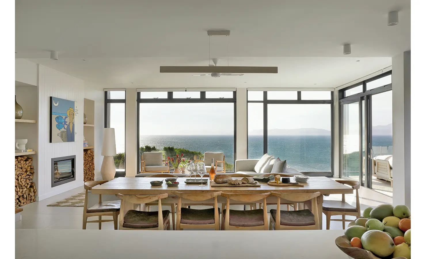Perfect Hideaways: A spacious, open-plan dining and lounge area with timber furniture and full-height windows framing breathtaking ocean views, seamlessly blending modern design with coastal tranquillity. Whales Breath, De Kelders.
