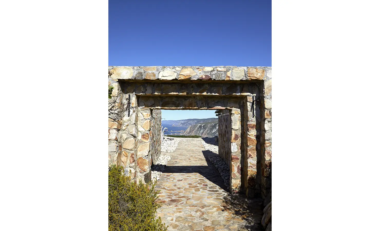 Perfect Hideaways: A rustic stone archway frames a cobbled path leading to sweeping coastal views, inviting guests into the natural drama of the landscape. Fynbos Fort, Plettenberg Bay.