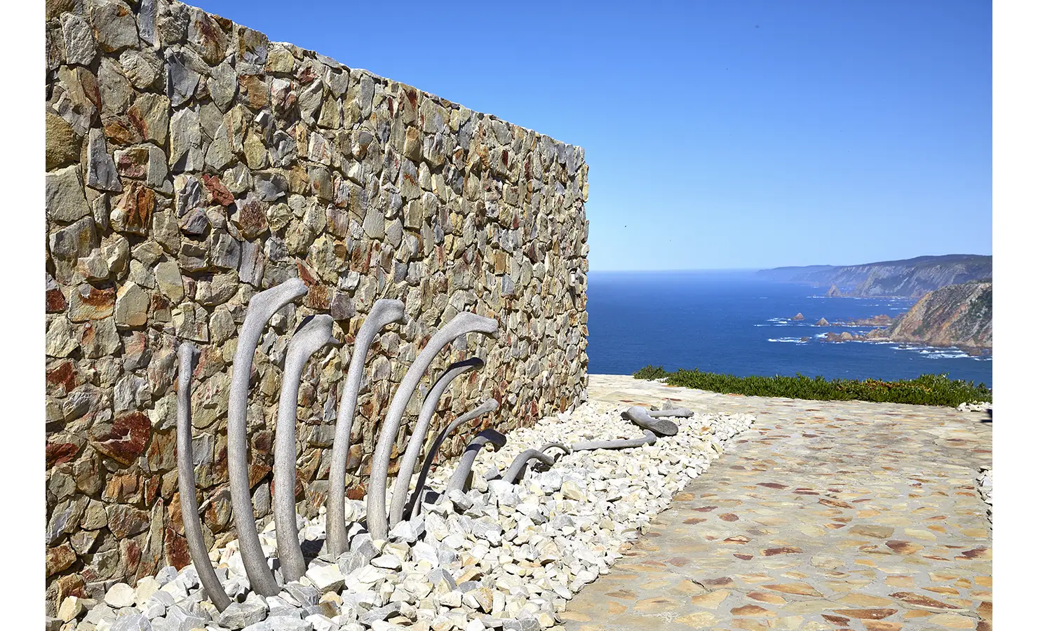 Perfect Hideaways: Curved driftwood bones rest against a rugged stone wall, set within a dry landscape garden, with panoramic sea views stretching beyond. Fynbos Fort, Plettenberg Bay.
