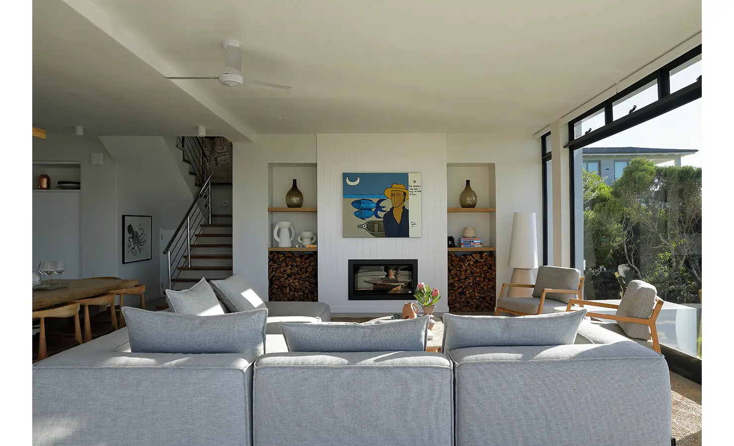 Perfect Hideaways: A cosy lounge with soft grey seating and modern fireplace flanked by shelves of firewood and curated ceramics, with large windows offering garden views and natural light. Whales Breath, De Kelders.