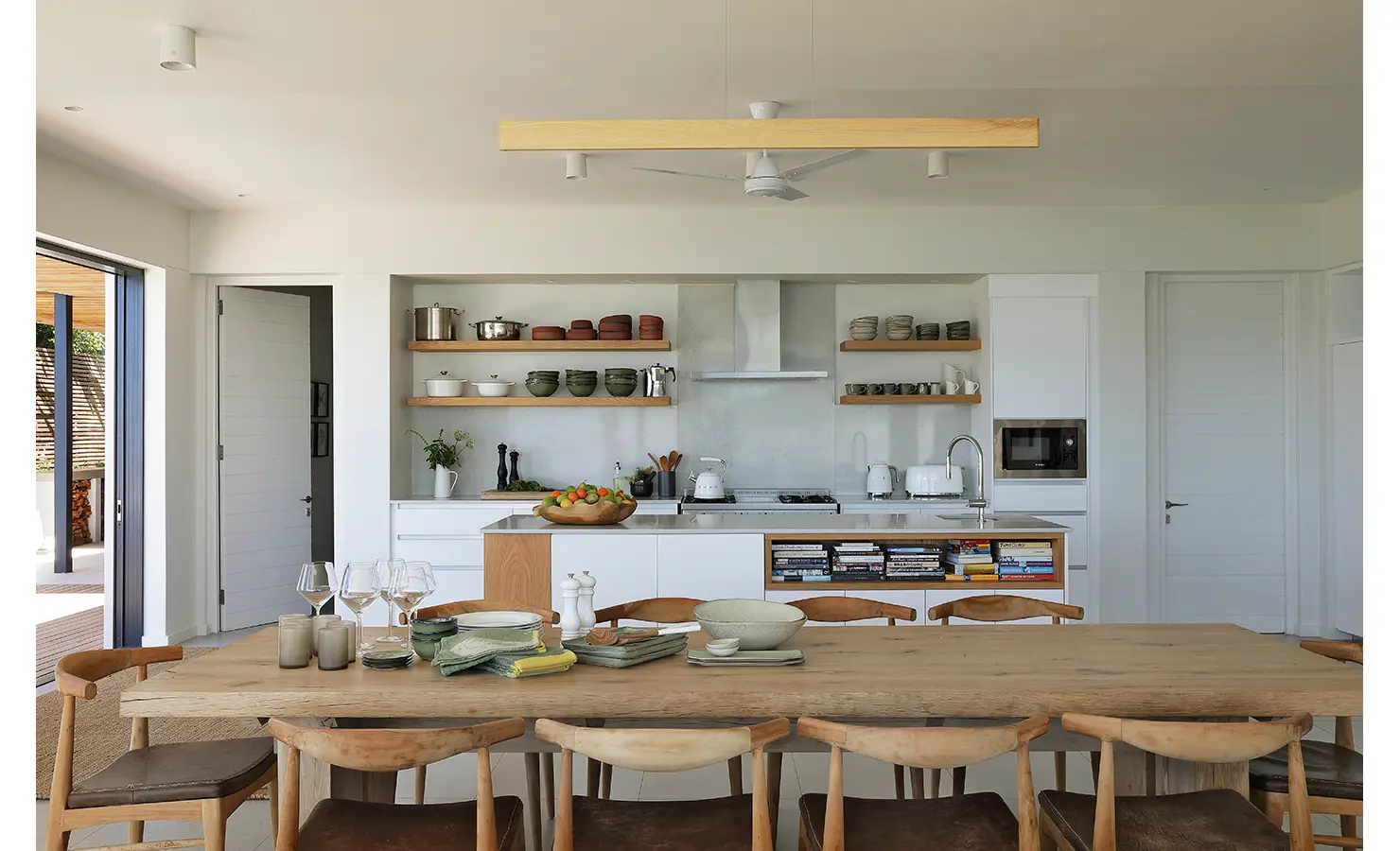 Perfect Hideaways: A chic open-plan kitchen with floating wooden shelves and curated ceramics, overlooking a generous timber dining table set with glassware and textiles, inviting relaxed, sociable meals. Whales Breath, De Kelders.