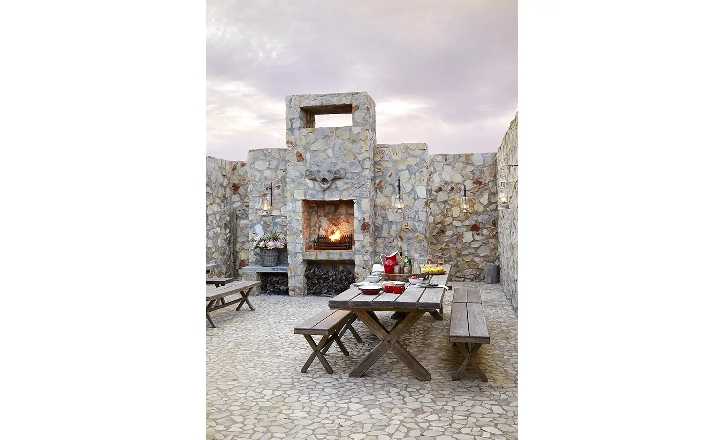 Perfect Hideaways: A dramatic stone courtyard with a glowing fire pit and long timber dining tables sets the scene for atmospheric al fresco gatherings. Fynbos Fort, Plettenberg Bay.