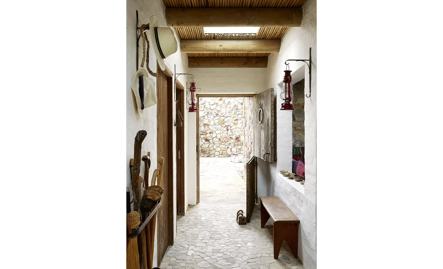 Perfect Hideaways: A charming passageway with hanging lanterns, vintage hats, and rustic textures leads toward a sunlit stone courtyard. Fynbos Fort, Plettenberg Bay.