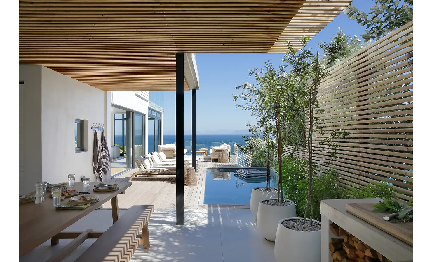 Perfect Hideaways: A shaded patio with slatted timber ceiling, outdoor dining area, and lush potted greenery leading to a sun-drenched deck and plunge pool with views of the Atlantic Ocean. Whales Breath, De Kelders.