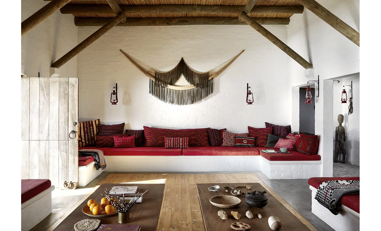 Perfect Hideaways: A cosy built-in lounge in warm reds and earthy tones is framed by exposed wooden beams and artisanal wall hangings, evoking relaxed, textured charm. Fynbos Fort, Plettenberg Bay.