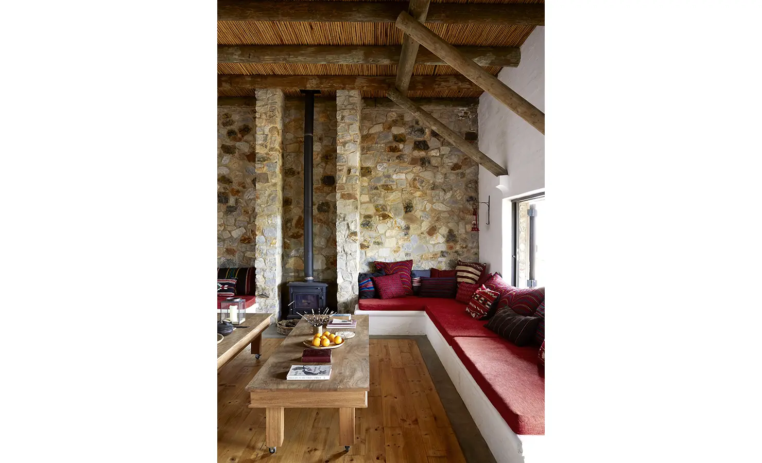 Perfect Hideaways: A character-filled living space with red-cushioned built-in seating and a wood-burning stove, set against stone walls and reed ceilings. Fynbos Fort, Plettenberg Bay.