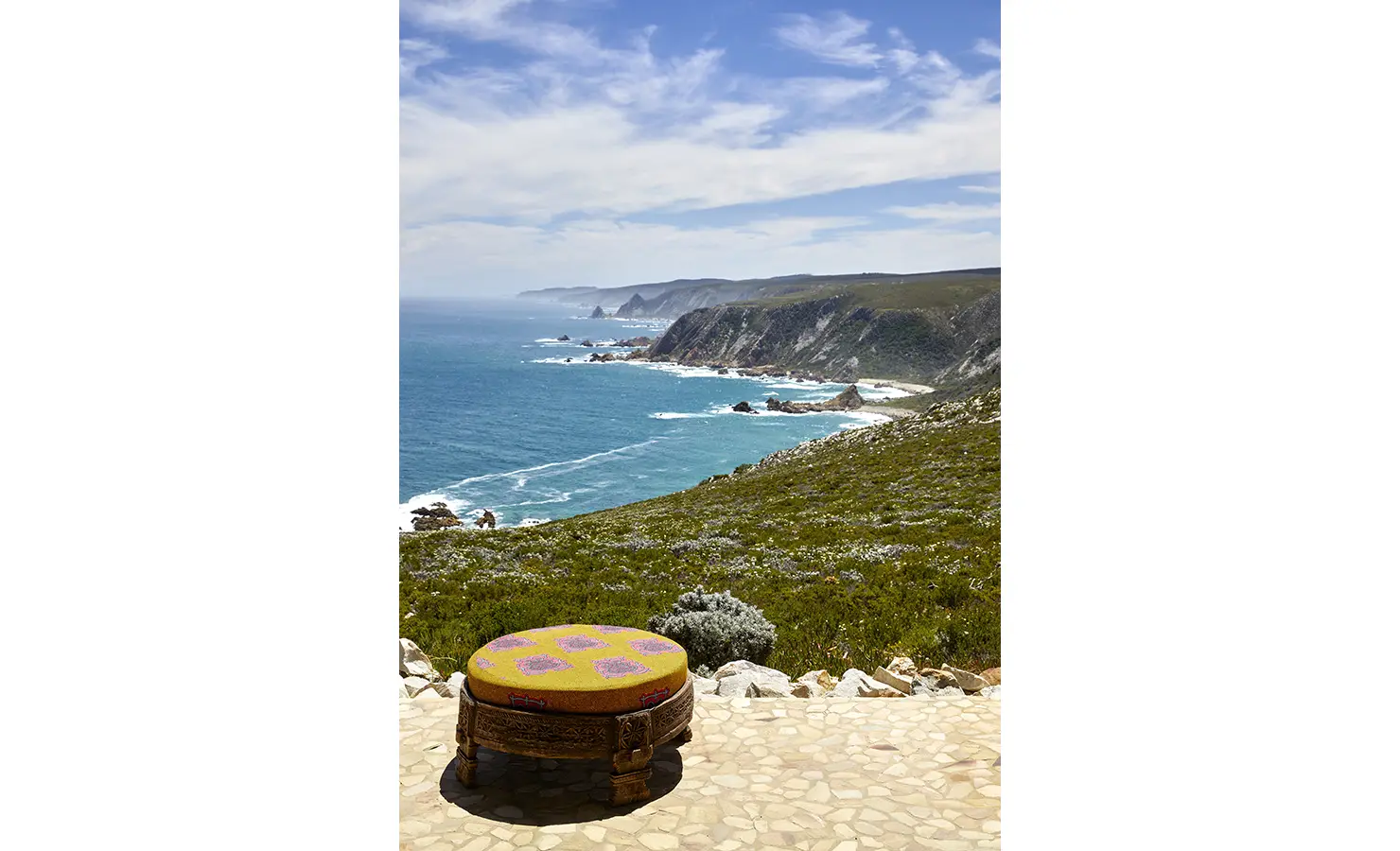 Perfect Hideaways: A vivid outlook across the wild coastline and turquoise ocean is anchored by a single colourful ottoman on a stone terrace. Fynbos Fort, Plettenberg Bay.