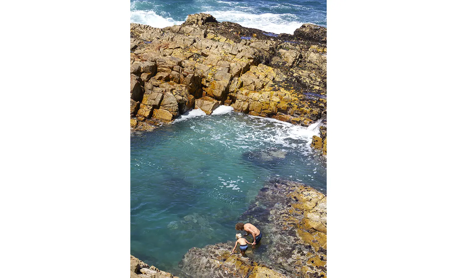 Perfect Hideaways: A father and child explore a secluded natural tidal pool nestled in golden rock formations, where turquoise waters meet the ocean's edge. Fynbos Fort, Plettenberg Bay.