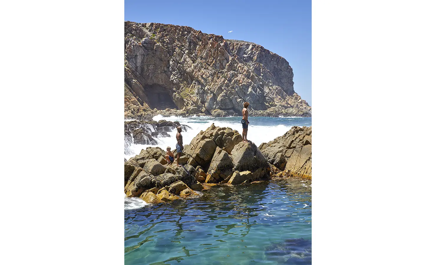 Perfect Hideaways: Children and adults perch on rugged coastal rocks beside crystal-clear tidal pools, with towering cliffs and a vivid blue sky behind. Fynbos Fort, Plettenberg Bay.