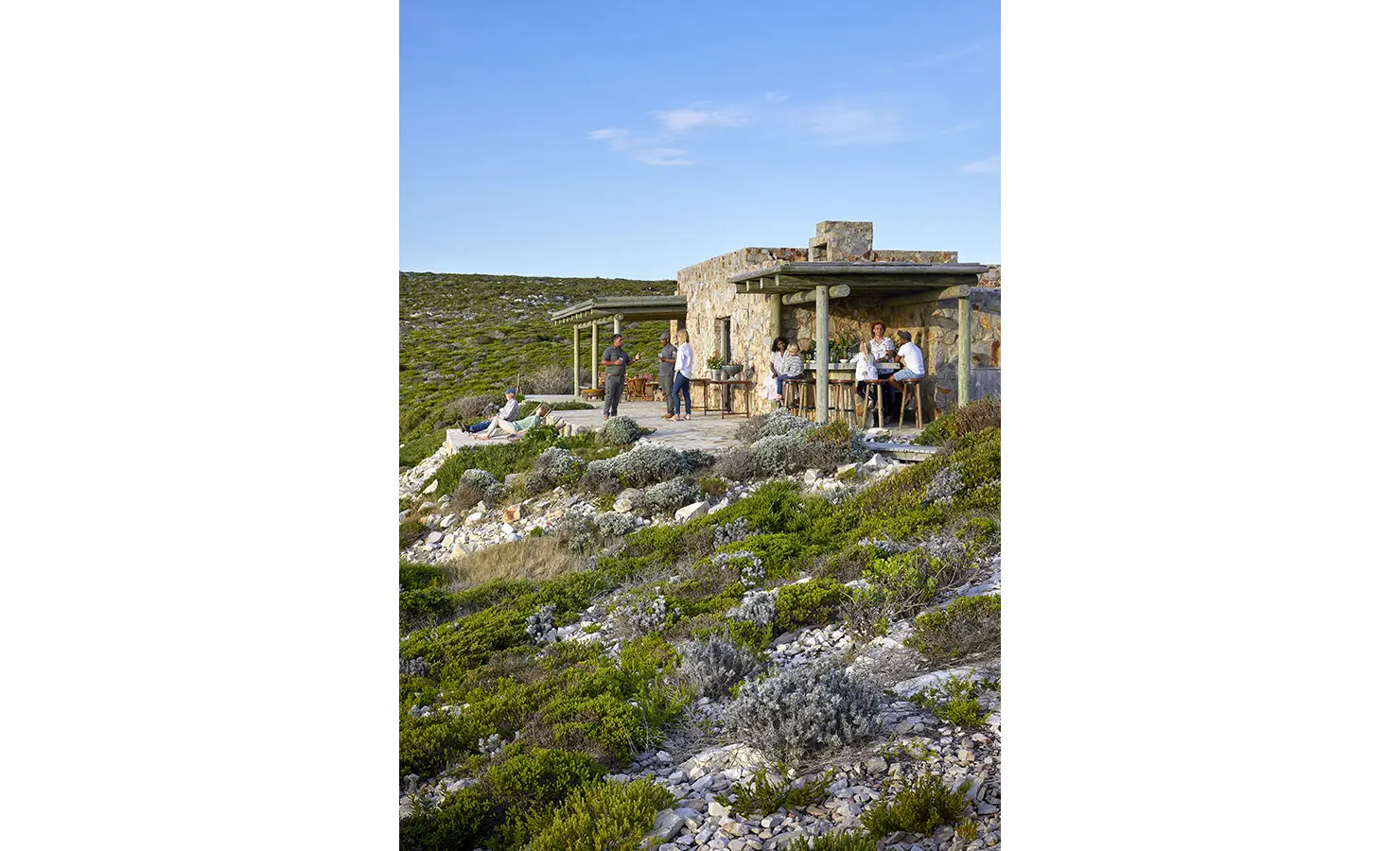 Perfect Hideaways: A group of friends gather on a wide stone terrace surrounded by fynbos, enjoying drinks and conversation at a rustic mountain retreat. Fynbos Fort, Plettenberg Bay.