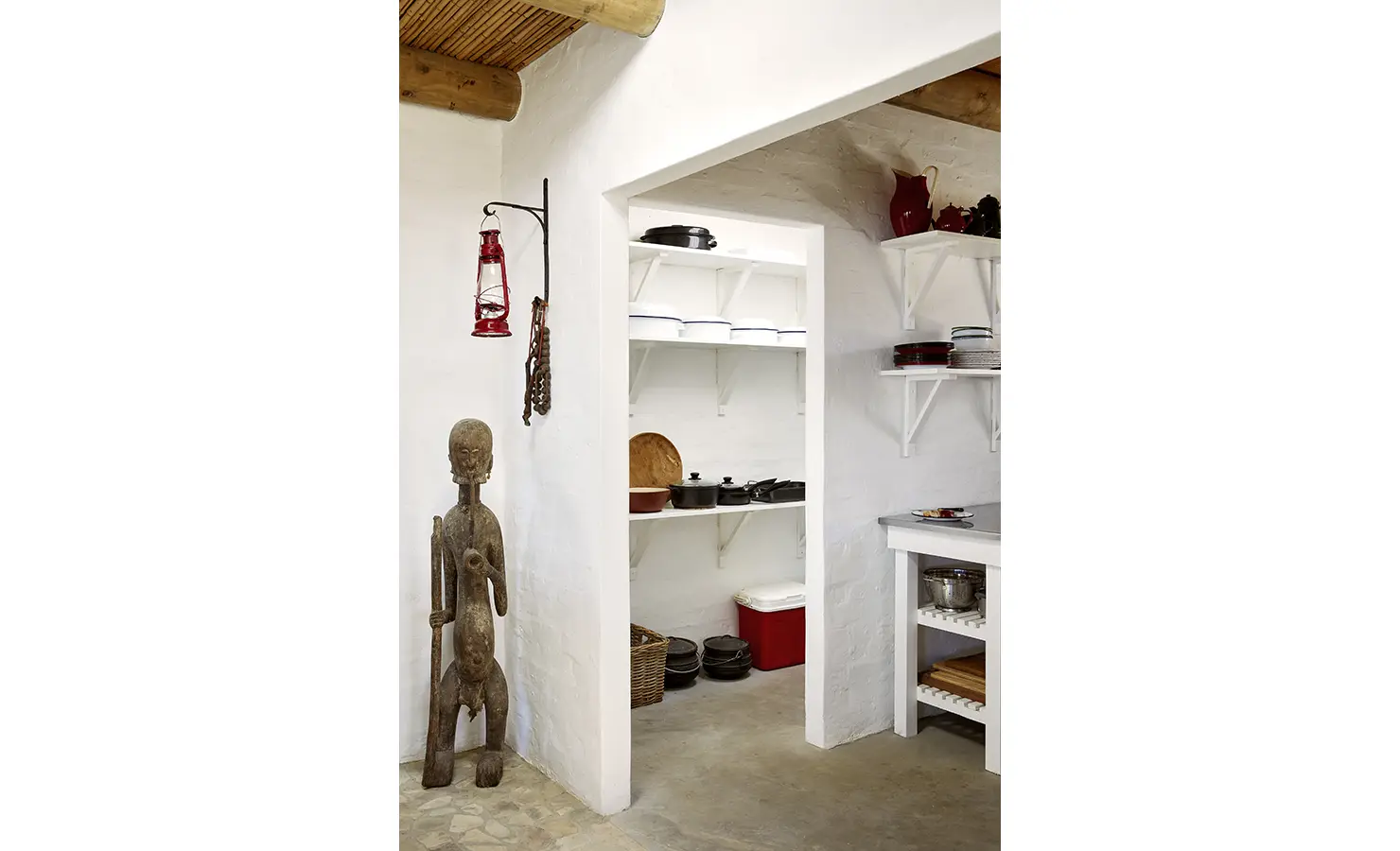 Perfect Hideaways: A minimalist scullery space with open shelving and vintage kitchenware is accented by a carved wooden statue and hanging lanterns. Fynbos Fort, Plettenberg Bay.