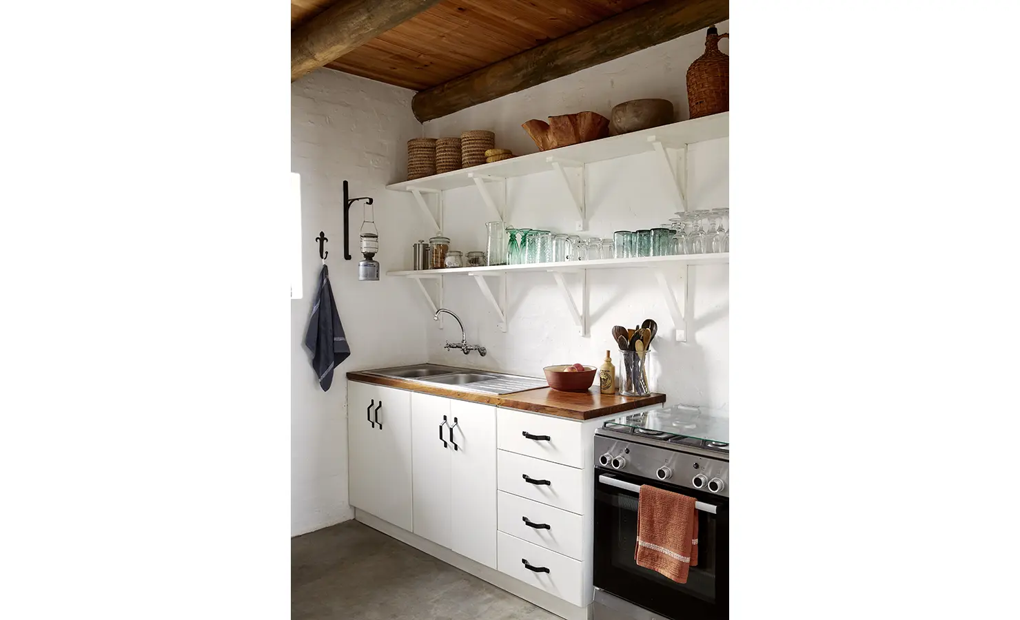Perfect Hideaways: A simple yet charming kitchen with white open shelving and a wooden countertop, warmed by woven baskets and antique vessels. Fynbos Fort, Plettenberg Bay.