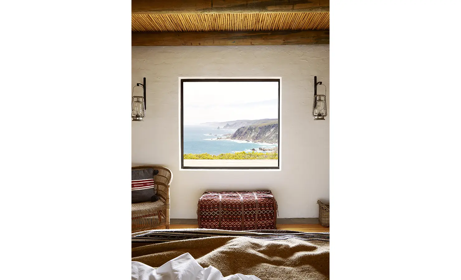 Perfect Hideaways: A framed sea view fills a bedroom window, with textured walls, lantern sconces and woven seating enhancing the natural ambience. Fynbos Fort, Plettenberg Bay.