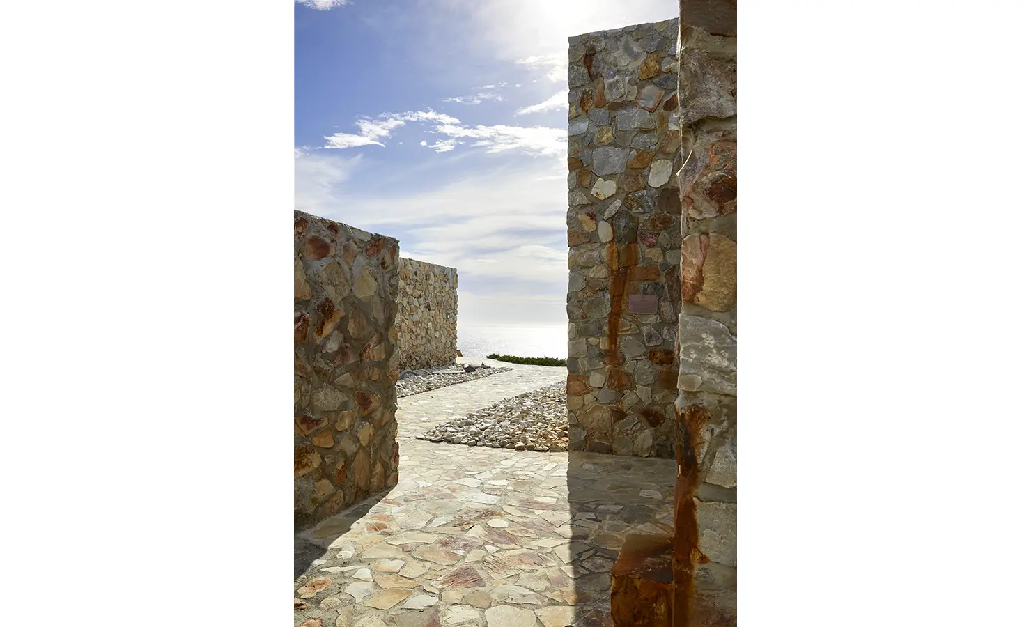 Perfect Hideaways: A sunlit stone walkway meanders between rugged stone walls, leading toward the vast ocean horizon under a cloud-dappled sky. Fynbos Fort, Plettenberg Bay.