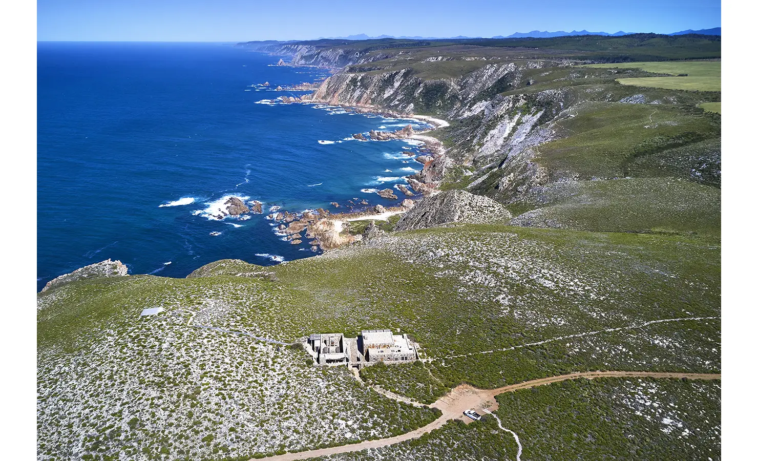Perfect Hideaways: An aerial shot captures the dramatic coastline and rugged landscape surrounding the secluded stone hideaway above the crashing waves. Fynbos Fort, Plettenberg Bay.