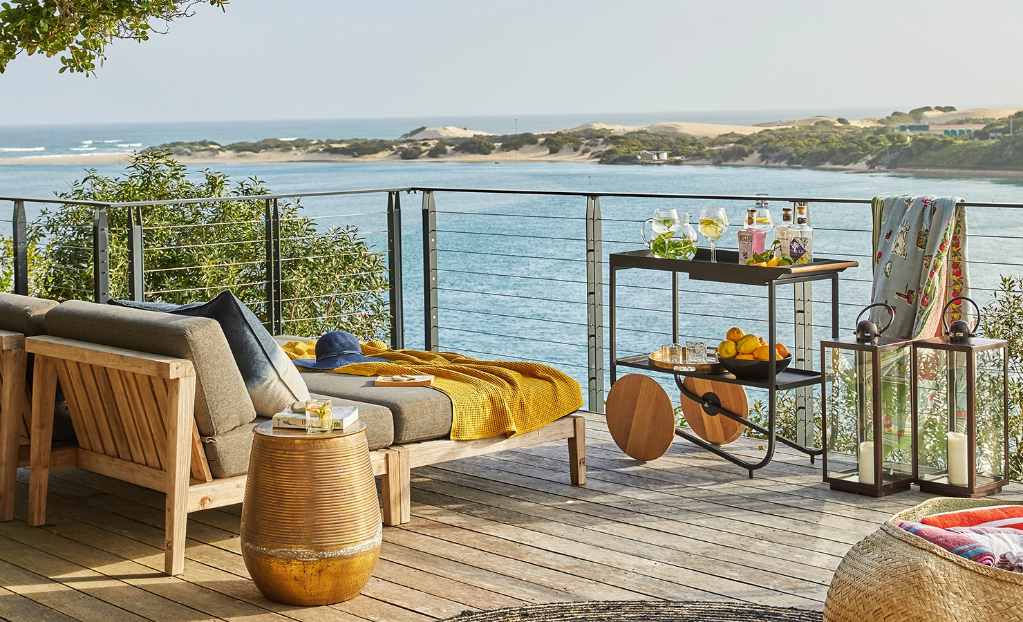 Perfect Hideaways: A stylish wooden deck with cushioned loungers, a golden side table, drinks trolley, and lanterns overlooks the sparkling estuary and sand dunes beyond, inviting relaxed outdoor living. St Austel, Kenton-on-Sea
