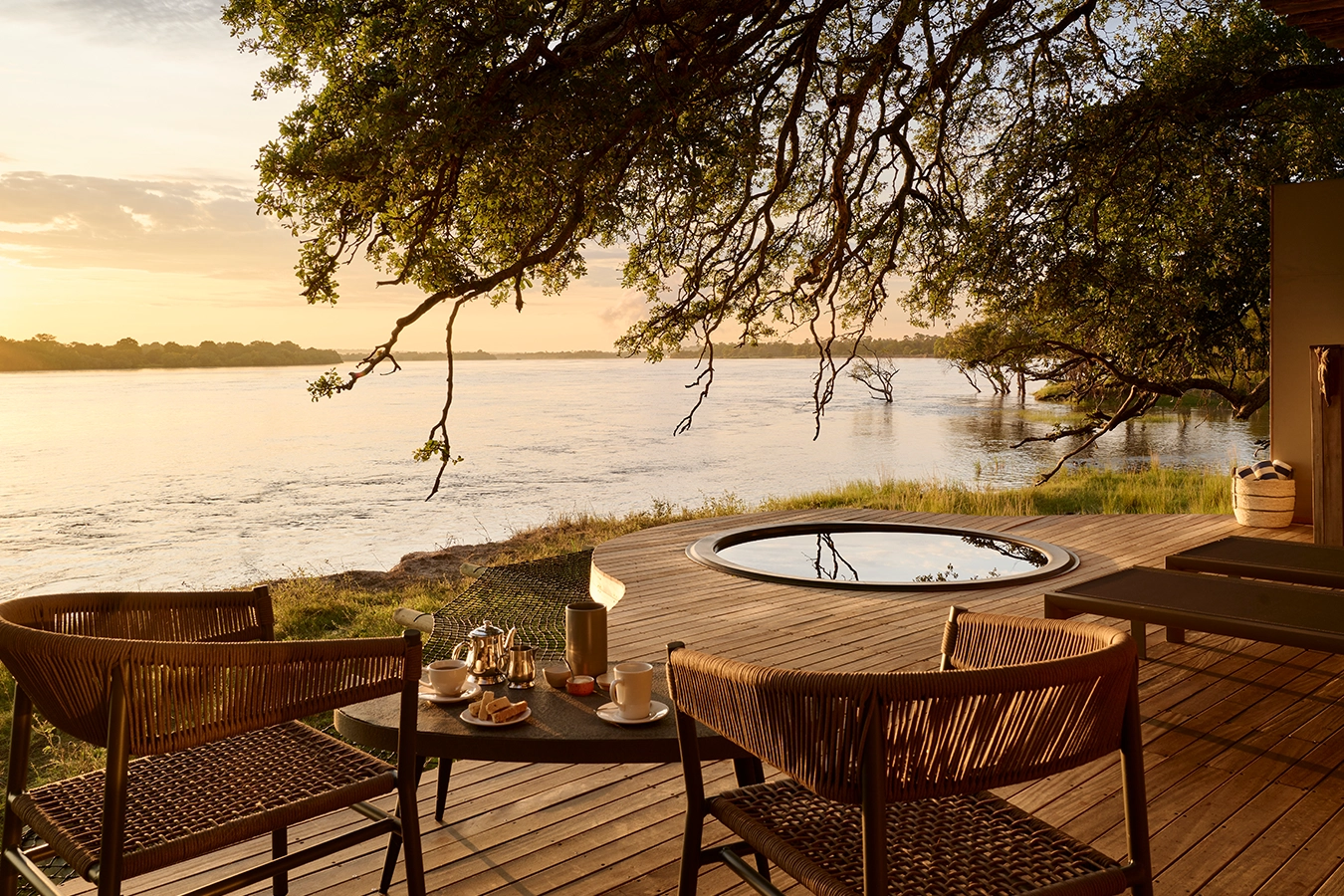 Perfect Hideaways: River-facing timber deck at golden hour with two woven chairs, breakfast tray and a circular plunge pool beneath overhanging branches, looking across the wide Zambezi. Hippo Creek Villa, Zambezi National Park.