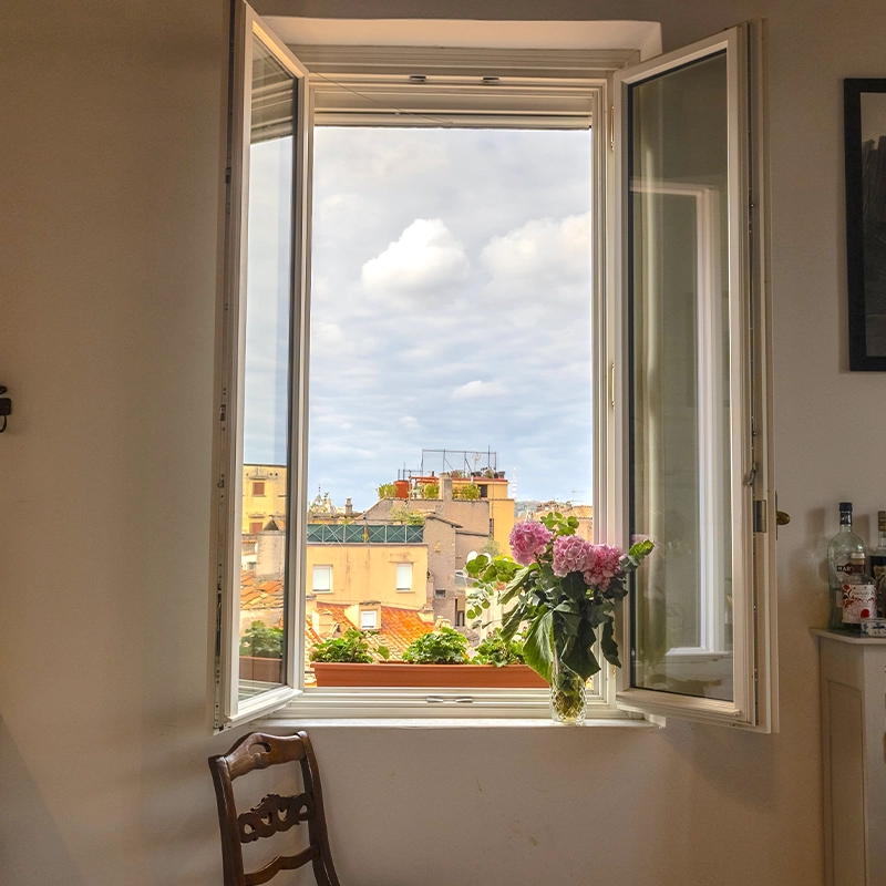 Perfect Hideaways: Sunlit kitchen with open casement windows framing Rome’s terracotta rooftops and soft clouds, a window box of greenery and a vase of pink hydrangeas on the sill, marble worktop and vintage drinks cabinet, Terracotta, Rome